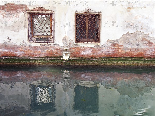 Two ornate windows with rusting iron bars in an ancient white plastered brick wall reflected in the still green water of a canal in venice italy