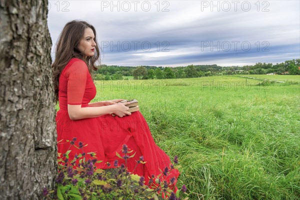 Beautiful woman, in an elegant red dress, sitting on a bench under a tree, with a book and pocket watch in her hands, in Schwabisch Hall, Germany, Europe