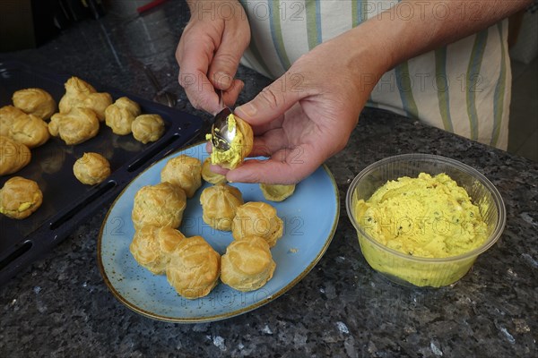 Swabian cuisine, preparing mini cream puffs, filling eclairs with cream cheese, profiterole, shortbread, oven cakes, choux pastry, pastry, hearty, salty, choux pastry, baked, from the oven, vegetarian, baking, typical Swabian reinterpreted, party pastry, finger food, appetisers for on the go, traditional cuisine reinterpreted, men's hands, plate, coffee spoon, fresh goat's cheese, baking tray, food photography, studio, Germany, Europe