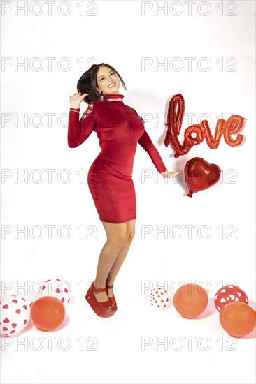 Young latina girl in red dress and shoes jumping funny, looks and smiles at camera, surrounded by balloons on white background with love sign. Valentine's day concept