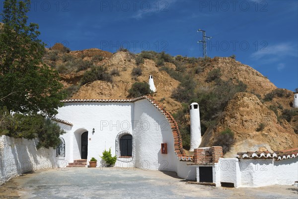 Small white building with chimney, tree and rock face in the background under a sunny blue sky, Pedro Antonio de Alarcon cave hotel, Guadix, Granada province, Andalusia, Spain, Europe