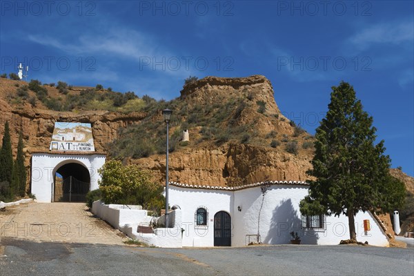 White building with entrance gate and trees in front of a rock face, under a sunny, cloudy sky, Pedro Antonio de Alarcon cave hotel, Guadix, Granada province, Andalusia, Spain, Europe