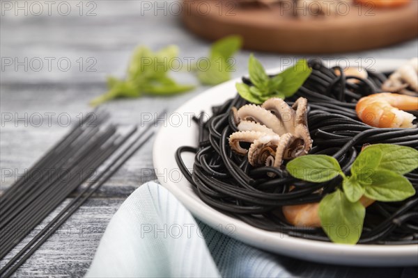 Black cuttlefish ink pasta with shrimps or prawns and small octopuses on gray wooden background and blue textile. Side view, close up, selective focus