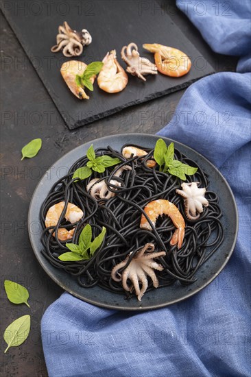 Black cuttlefish ink pasta with shrimps or prawns and small octopuses on black concrete background and blue textile. Side view, close up