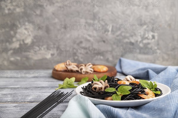 Black cuttlefish ink pasta with shrimps or prawns and small octopuses on gray wooden background and blue textile. Side view, copy space, selective focus