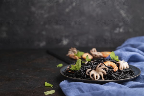 Black cuttlefish ink pasta with shrimps or prawns and small octopuses on black concrete background and blue textile. Side view, copy space, selective focus