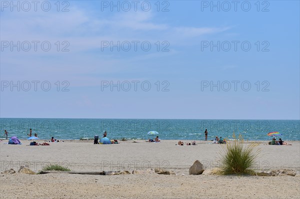 Beach with people and colourful umbrellas, stones and grass, in front of a wide, blue Mediterranean Sea under a clear sky, Saintes-Maries-de-la-Mer, Camargue, France, Europe