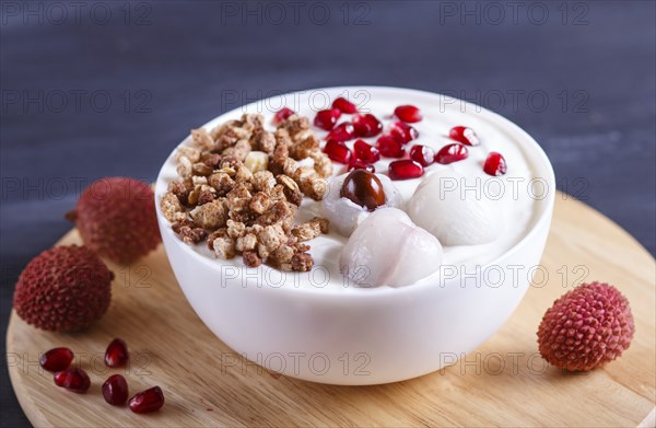 Greek yogurt with lychee, pomegranate seeds and granola in a white plate on a black wooden background, close up