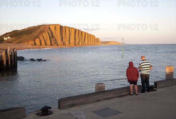 Golden afternoon light on sandstone cliffs, East Cliffs, West Bay, Bridport, Dorset, England, UK with father and son fishing in the foreground