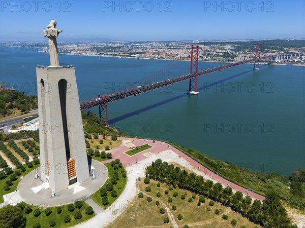 A tall statue with outstretched arms stands on a viewpoint near a bridge on the water, with a city in the background, aerial view, Statue Cristo Rei, Almada, Ponte 25 de Abril, 25 April Bridge, suspension bridge over the Tagus, double-decker bridge, road traffic and railway traffic, Lisbon, Lisboa, Portugal, Europe