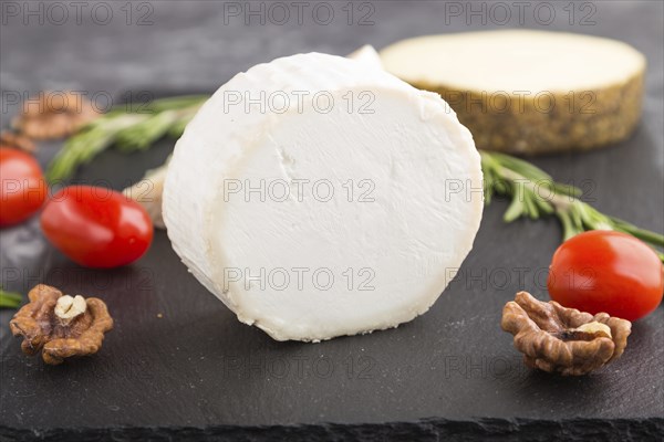 White goat cheese and various types of cheese with rosemary and tomatoes on black slate board on a black concrete background. Side view, close up, selective focus