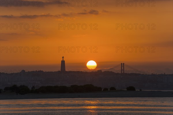 A sunset over a city by the sea with a striking statue and a bridge in the background, Statue Cristo Rei, Ponte 25 de Abril, Bridge of 25 April, Suspension bridge over the Tagus, Lisbon, Lisboa, Portugal, Europe