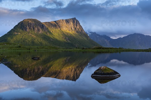 Landscape on the Lofoten Islands. Vagspollen bay, with Offersoykammen mountain behind it. The mountain Stornappstinden on Flakstadoya in the background. The mountains are reflected in the water. At night at the time of the midnight sun. A few clouds in the sky. The night sun shines on the mountain peak. Early summer. Golden hour. Vestvagoya, Lofoten, Norway, Europe