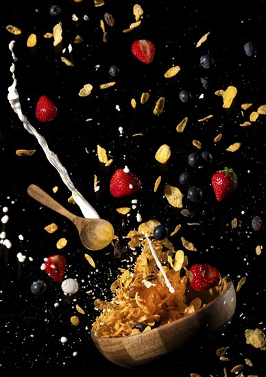 Cornflakes, milk, strawberries and blueberries fly out of a bowl against a black background