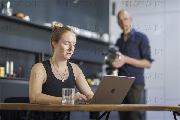 Symbolic photo on the subject of division of labour for couples in the household. A woman sits at a laptop in a kitchen while a man washes dishes in the background. Berlin, 13.08.2024