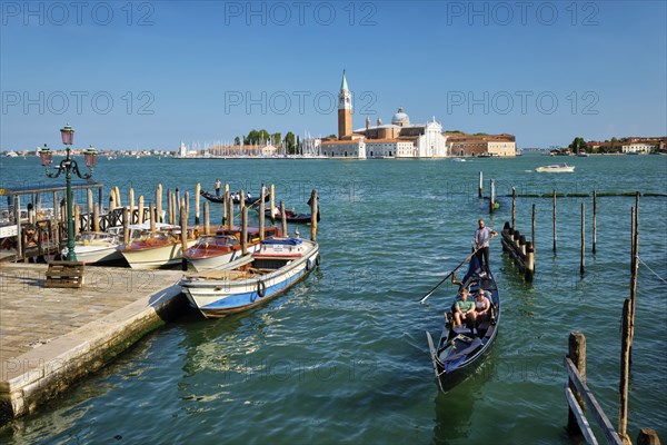 VENICE, ITALY, JULY 19, 2019: Gondolier with tourists in gondola in lagoon of Venice by Saint Mark San Marco square San Giorgio di Maggiore church in Venice, Italy seen from Ponte della Paglia