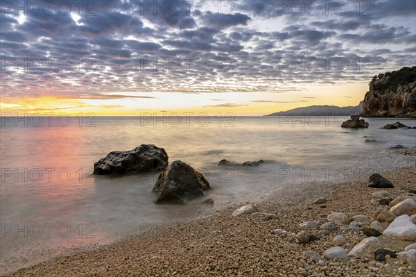 A colorful sunrise at Cala Gonone beach with black and white rocks and boulders in the foreground