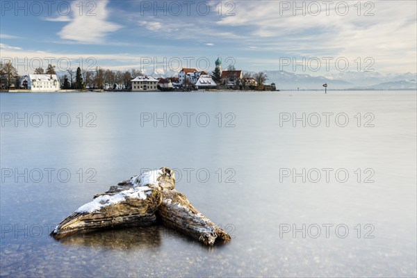 A peaceful lake with a log in the foreground with picturesque St George's Church on the lakeshore in front of snow-covered Pfänder on a clear winter's day, moated castle, Lake Constance, Bavaria, Germany, Europe