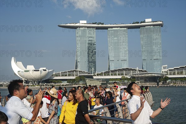 01.08.2019, Singapore, Republic of Singapore, Asia, Tourists pose for photos in Merlion Park on the banks of the Singapore River with the Marina Bay Sands Hotel in the background. The open mouth is meant to catch the stream of water from the Merlion figure, which is not in the picture, Asia