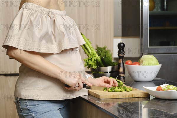 Closeup view of hands of pregnant unrecognizable woman standing in profile in the kitchen cutting bell pepper for salad