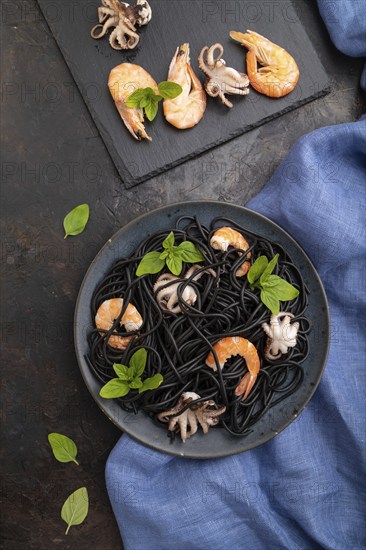 Black cuttlefish ink pasta with shrimps or prawns and small octopuses on black concrete background and blue textile. Top view, flat lay, close up