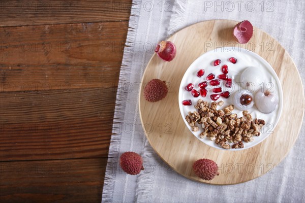 Greek yogurt with lychee, pomegranate seeds and granola in a white plate on a brown wooden background, top view, flat lay, copy space, selective focus