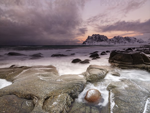 Rounded rocks on Utakleiv beach in a dramatic cloudy atmosphere, snow-capped mountains in the background, Vestvagoy Island, Lofoten, Norway, Europe