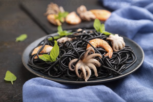 Black cuttlefish ink pasta with shrimps or prawns and small octopuses on black concrete background and blue textile. Side view, close up, selective focus