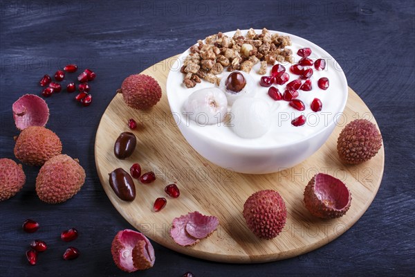 Greek yogurt with lychee, pomegranate seeds and granola in a white plate on a black wooden background, close up