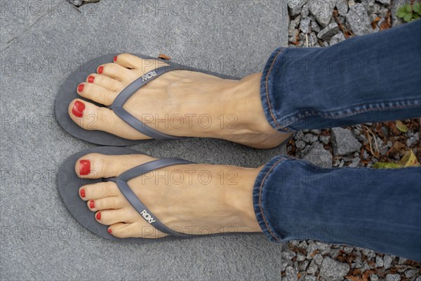 Feet of a woman, mid-50s, toes painted red, wearing jeans trousers, sandals, flip-flops