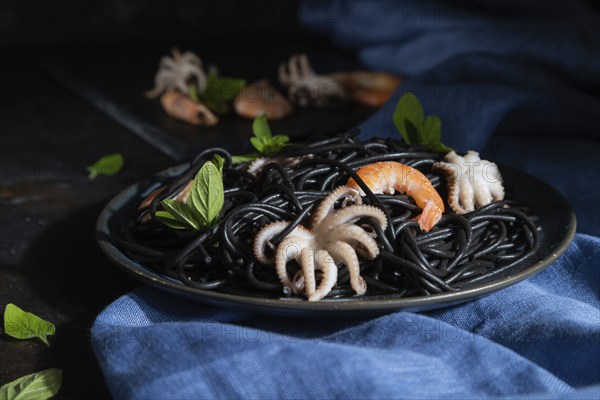 Black cuttlefish ink pasta with shrimps or prawns and small octopuses on black concrete background and blue textile. Low key, hard light. Side view, close up