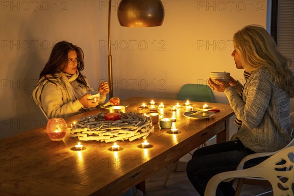 Symbolic image energy saving, cold winter, energy crisis, cold flat, mother and daughter dressed warmly at dinner, only by candlelight, heating on to a minimum