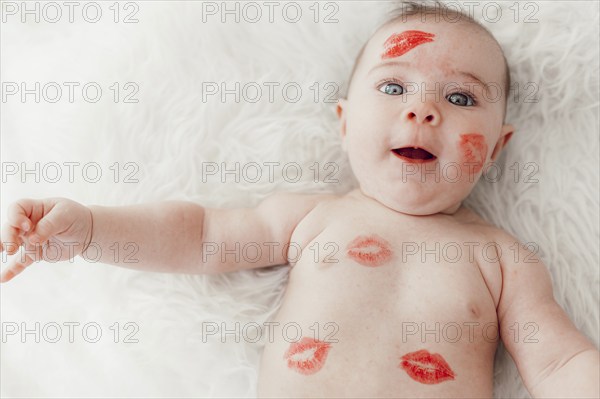 A smiling baby with red lipstick kisses on her face and body lies on a white blanket and looks into the camera