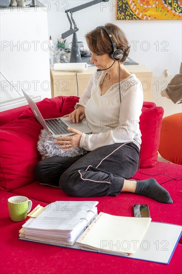 Woman, mid-50s, works from home, with laptop and communicates with colleagues via headset, home office, on the sofa