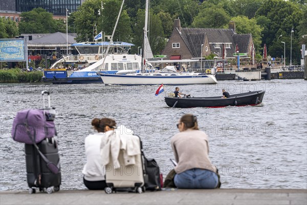 Tourists sitting at the back of Amsterdam Centraal railway station, on the banks of the river Ij, Amsterdam, Netherlands