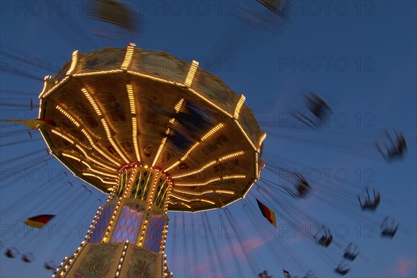 An illuminated carousel at dusk, spinning fast, in front of a blue sky with clouds and a Germany flag, Killiani Volksfest, Würzburg, Bavaria