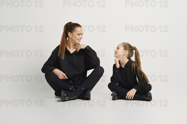 Mother and her daughter are sitting on a white background wearing similar stylish black shirt, tights and boots. They look at each other with smile