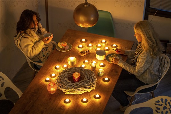 Symbolic image energy saving, cold winter, energy crisis, cold flat, mother and daughter dressed warmly at dinner, only by candlelight, heating on to a minimum