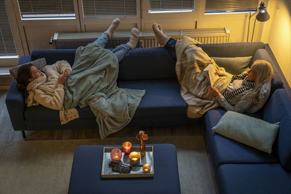 Symbolic image energy saving, cold winter, energy crisis, cold flat, mother and daughter lying on the sofa, warmly dressed, candlelight, heating on to a minimum, warming their feet on the radiator