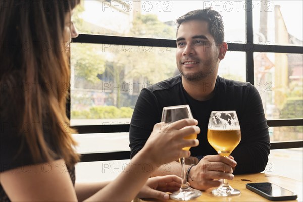 A young couple enjoys a craft beer at a bar during the day. They smile and engage in conversation, creating a warm and relaxed atmosphere. The bar has large windows overlooking the outside