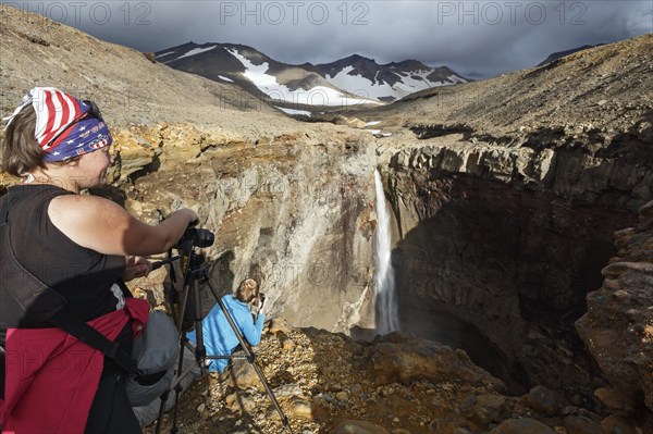 MUTNOVSKY VOLCANO, KAMCHATKA PENINSULA, RUSSIA, SEP 8, 2015: Young women tourists photographers takes picture of waterfall on mountain river in Dangerous Canyon with rocky slope on active volcano Mutnovskaya Sopka