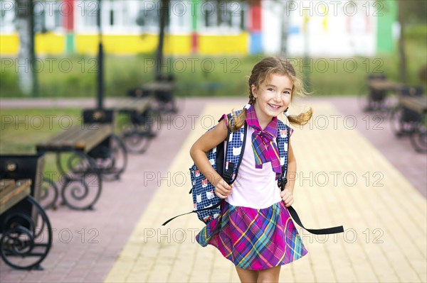 Cheerful girl with a backpack and in a school uniform in the school yard. Back to school, September 1. A happy pupil. Primary education, elementary class. Portrait of a student