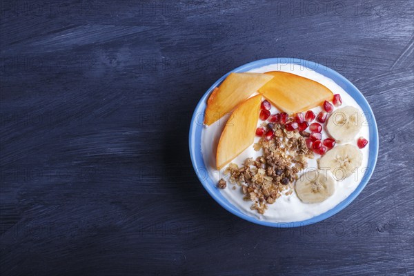 A blue plate with greek yogurt, granola, persimmon, banana, pomegranate black wooden background. top view. copy space