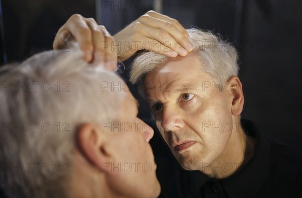 Symbolic photo on the subject of a high hairline and receding hairline. A man looks at his hairline in the mirror. Berlin, 13.08.2024