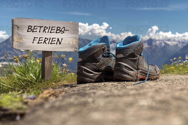 Symbolic image of company holidays/summer holidays: Hiking boots in front of an alpine backdrop and a sign with the inscription BETRIEBSFERIEN (company holidays)