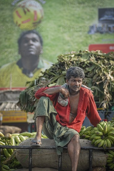 Worker on a cart with banana plants in front of a football poster, Thrissur, South India