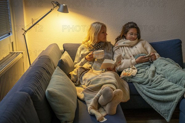 Symbolic image energy saving, cold winter, energy crisis, cold flat, mother and daughter lying on the sofa, warmly dressed, candlelight, heating on to a minimum