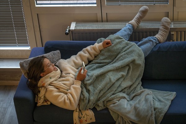 Symbolic image energy saving, cold winter, energy crisis, cold flat, mother and daughter lying on the sofa, warmly dressed, candlelight, heating on to a minimum, warming their feet on the radiator