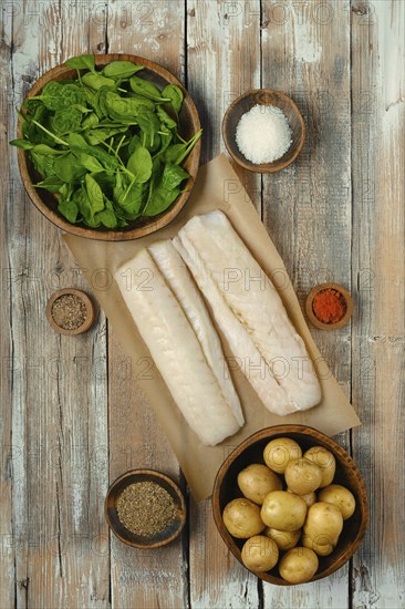 Fresh fish fillets on parchment paper surrounded by bowls of spinach, potatoes, and various spices, ready for meal preparation on a rustic wooden table, perfect for cooking enthusiasts