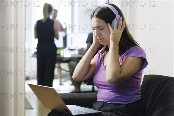 Topic: A woman works with headphones on her laptop while colleagues talk loudly. Berlin, 08.08.2024